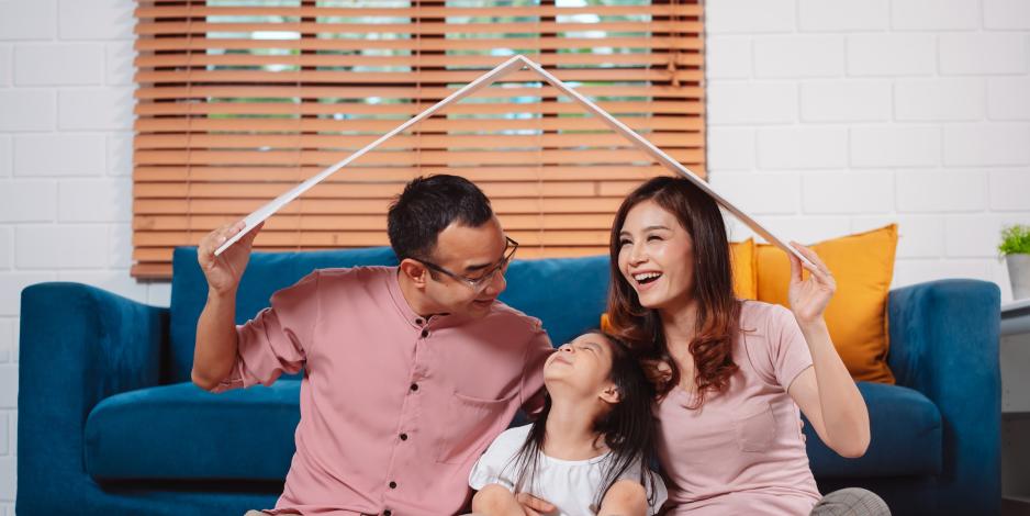 Happy family holding a model roof over their head as they sit and laugh in a living room Happy family holding a model roof over their head as they sit and laugh in a living room